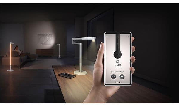 Lightcycle Morph Floor:  Kvinna som sitter i en soffa medan en hand i förgrunden håller en telefon med Dyson-appen öppen.