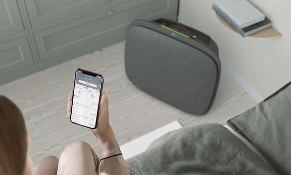 En mørkegrå Well 5 på gulvet og en kvinne med Wellbeing-appen på telefonen