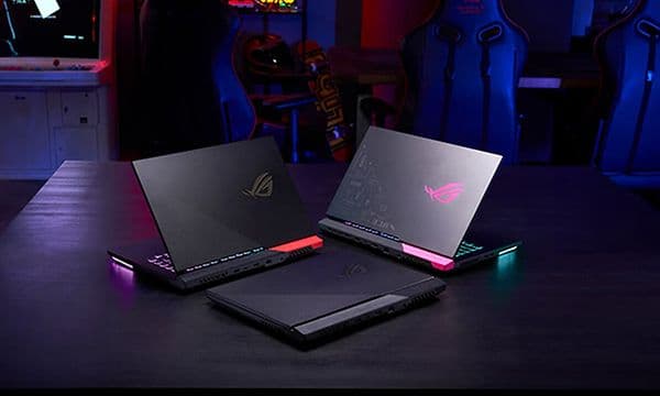Tre ROG Strix G15 bærbar gaming-PCer oppstilt på et bord med gamingstoler og utstyr i bakgrunnen.