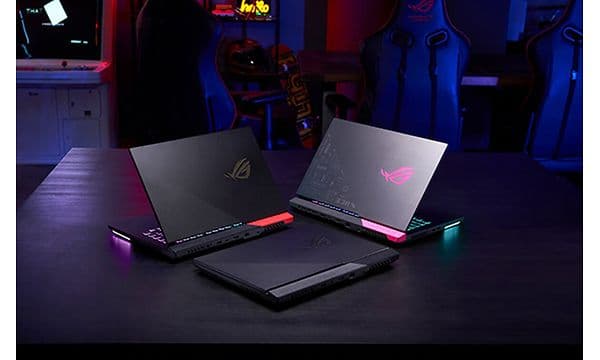 Tre ROG Strix G15 bærbar gaming-PCer oppstilt på et bord med gamingstoler og utstyr i bakgrunnen.