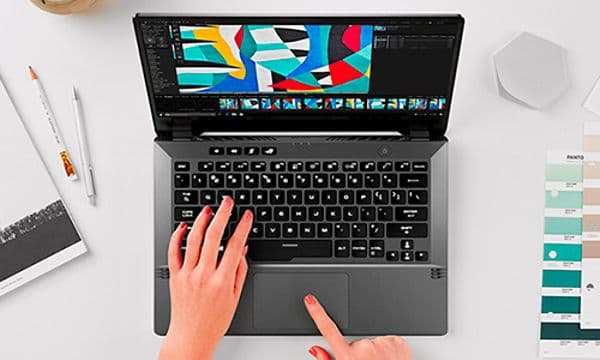 En designer jobber på en ROG Zephyrus G14, med fargeprøver plassert på pulten og et fargerikt motiv på skjermen.