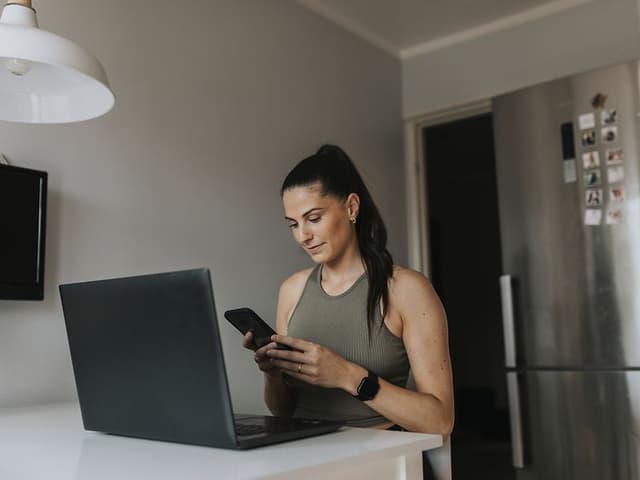 En kvinne leser på smarttelefonen sin foran en laptop på kjøkkenet