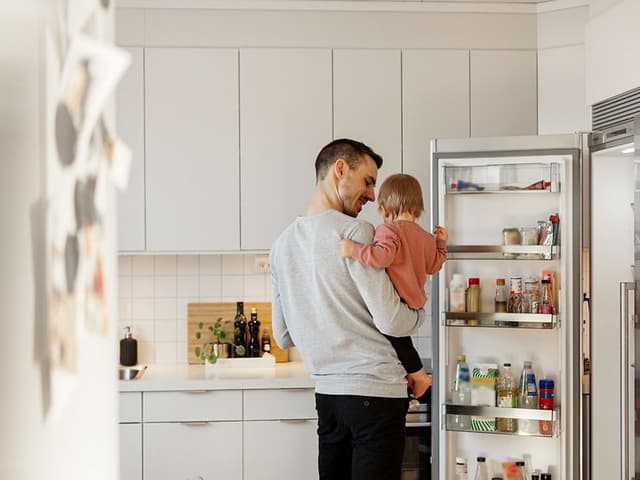 En mann med et barn på armen ser inn i et kjøleskap