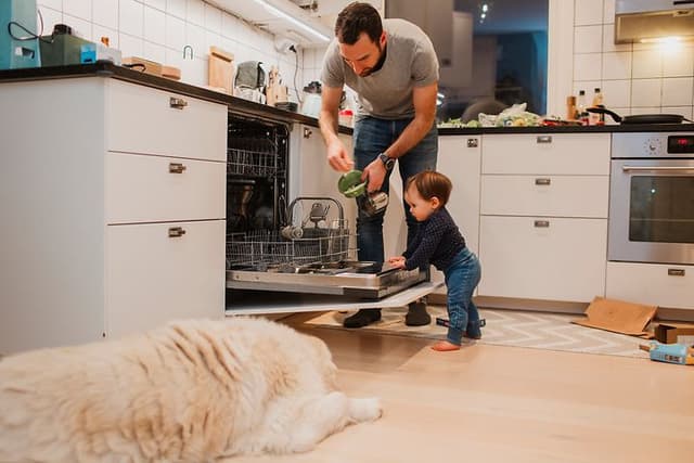 En mann og et barn legger inn skitne tallerkner i oppvaskmaskinen og en hund ligger på gulvet