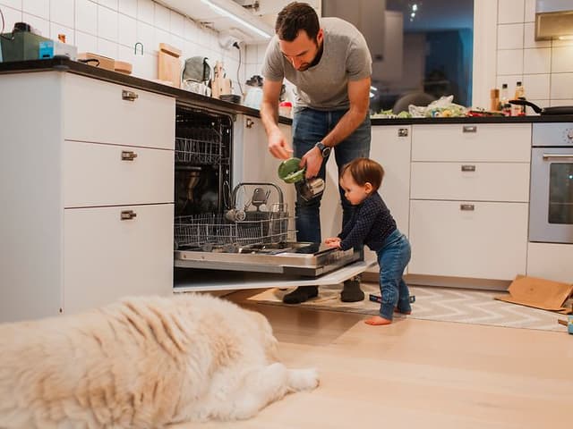 En mann og et barn legger inn skitne tallerkner i oppvaskmaskinen og en hund ligger på gulvet
