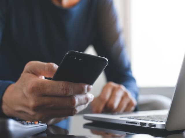 Nærbilde av en person som holder en smarttelefon foran en bærbar PC