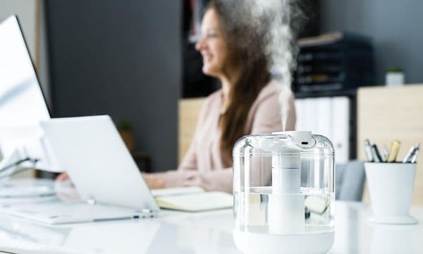En luftfuktare i ett kontor med en kvinna som ler och sitter vid ett skrivbord i bakgrunden 