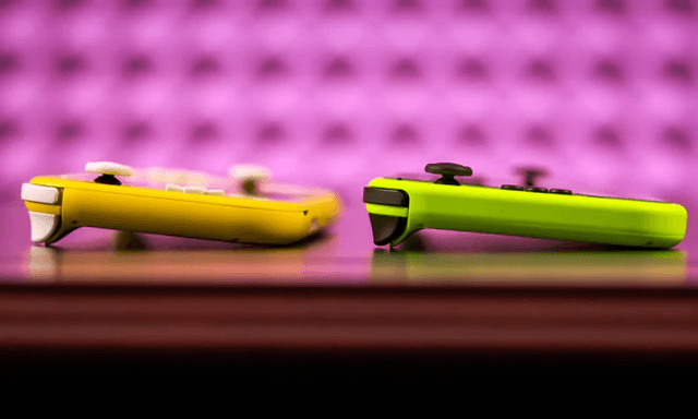Nintendo Switch Lite, två spelkonsoler på ett bord, en i beige färg och den andra grön. Rosa vägg i bakgrunden.