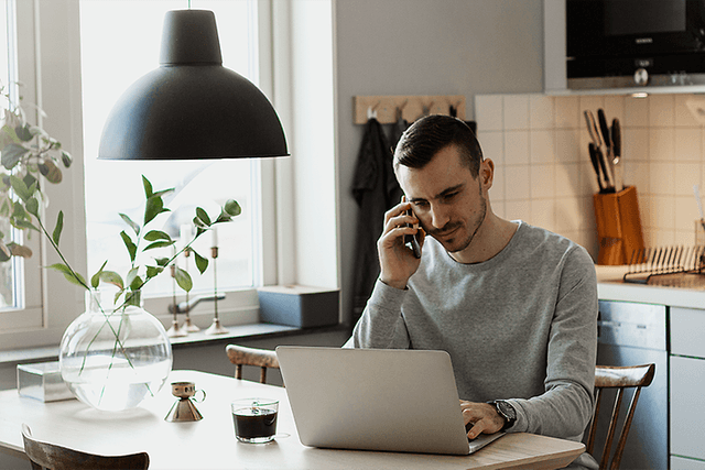 En mand, der arbejder på sin bærbare computer og taler på sin smartphone i et køkken