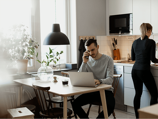 En mand, der arbejder på sin bærbare computer og taler på sin smartphone i et køkken