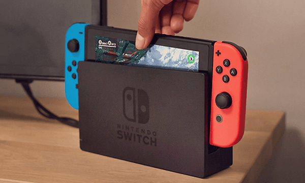 En hand placerar Nintendo Switch i en dockningsstation som är ansluten till en TV som syns i bakgrunden. 