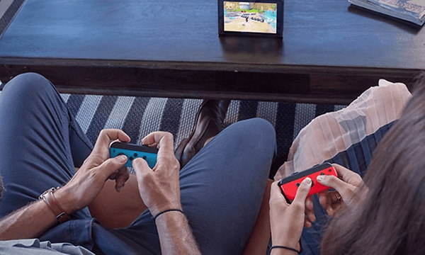 Bil ovanifrån på två personer som spelar Nintendo Switch med joycon-kontroller i händerna och bildskärm på bordet framför.  