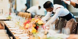En man i vit skjorta dukar upp ett långbord med glas, tallrikar och bestick. På bordet står även blombuketter. 