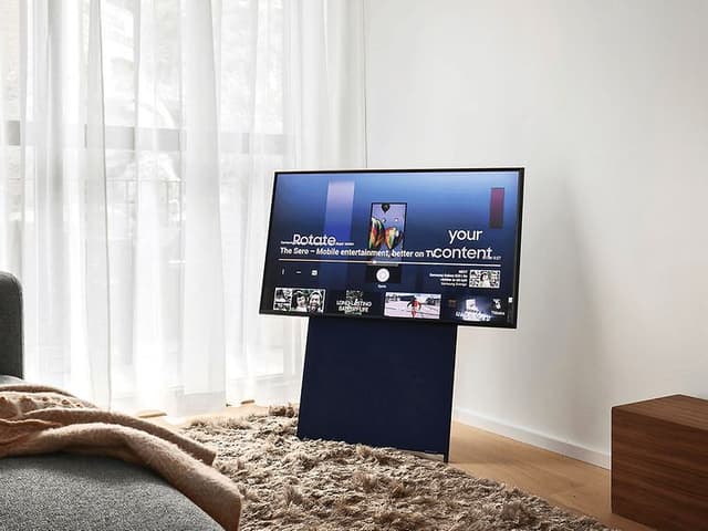 Samsung The Sero på stativ i vardagsrum med stora fönster, ljusa gardiner och en grå soffa.
