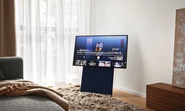Samsung The Sero på stativ i vardagsrum med stora fönster, ljusa gardiner och en grå soffa. 
