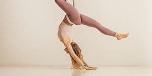 Kvinna tränar aerialyoga hängades i ett beigefärgat tyg från taket. Hon har ljusrosa linne och mörkrosa yogataights. 