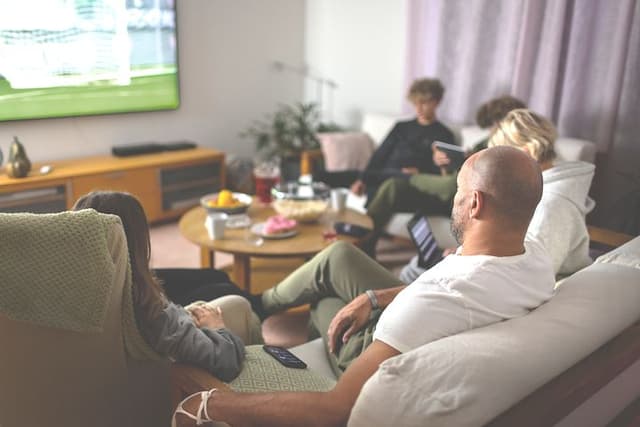 Familj i soffa som tittar på sport på TV och har snacks på ett runt bord i mitten