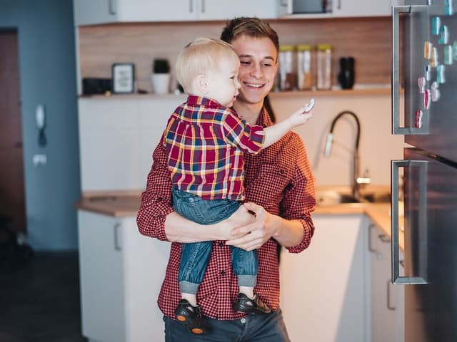 Isä pitelee pientä poikaansa sylissään, joka tutkii jääkaapin oveen kiinnitettyjä magneetteja
