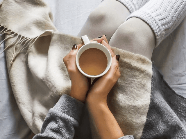 kvinne sitter i en seng med teppe over beina og kaffekopp i hendene