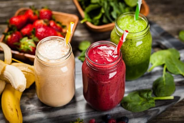 Tre glass med ulike smoothier med frukt og grønt liggende rundt