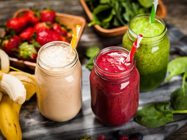 Tre glass med ulike smoothier med frukt og grønt liggende rundt
