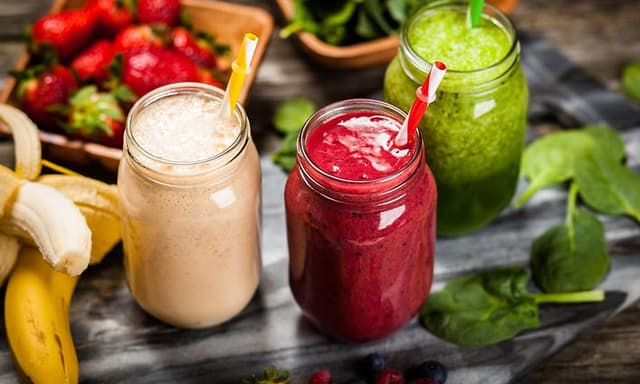 Tre glass med ulike smoothier med frukt og grønt liggende rundt