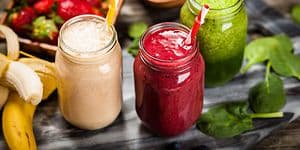 Tre glass med ulike smoothier med frukt og grønt liggende rundt