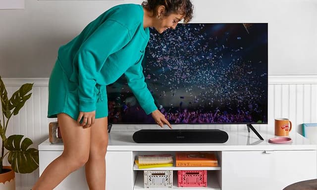 Glad kvinna i fritidskläder och tofflor trycker på en svart soundbar under en TV