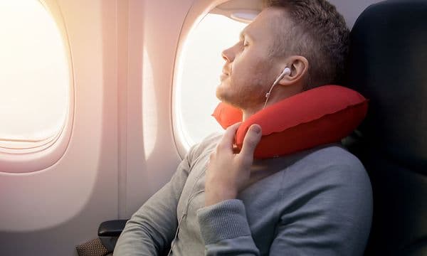 En mann sitter på et fly ved inntil et vindu og sover med en rød nakkepute
