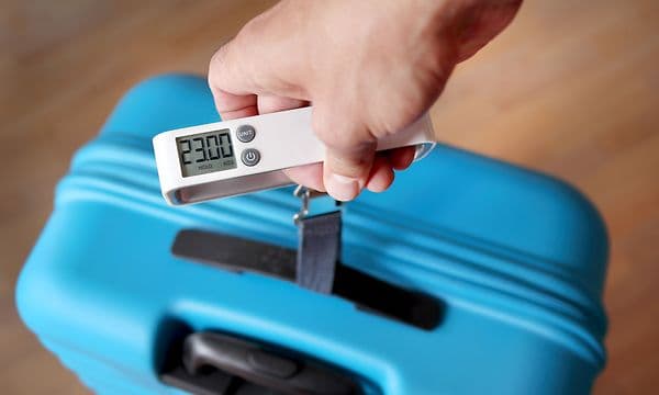 En hånd veier en blå koffert med en bagasjevekt