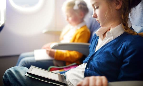 En ung jente sitter i et fly og leser på et lesebrett ved siden av en liten jente