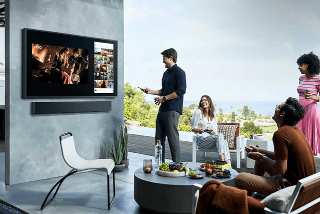 Samsung-Fyra personer tittar på The Terrace TV utomhus på en terrass med frukt på ett bord och med sol och utsikt över landskap