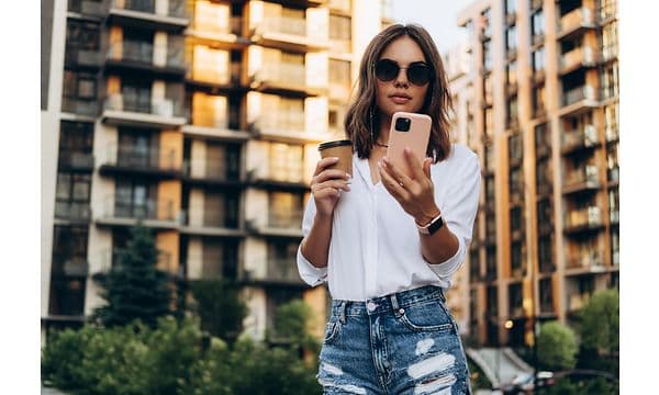 Kvinne med solbriller holder en kaffekopp og bruker en smarttelefon