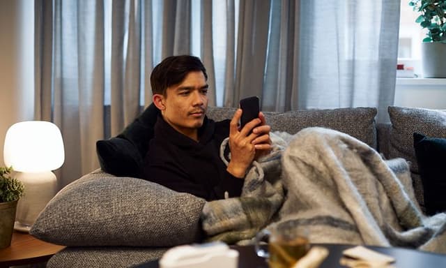 mann sitter i sofa med smarttelefon i hendene og smarte lamper i bakgrunnen