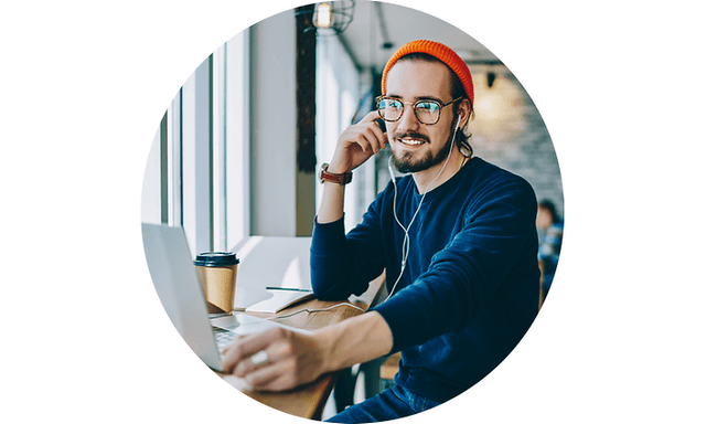 Glad man på café med mössa och hörlurar och en kaffemugg bredvid sig tittar på sin bärbara dator
