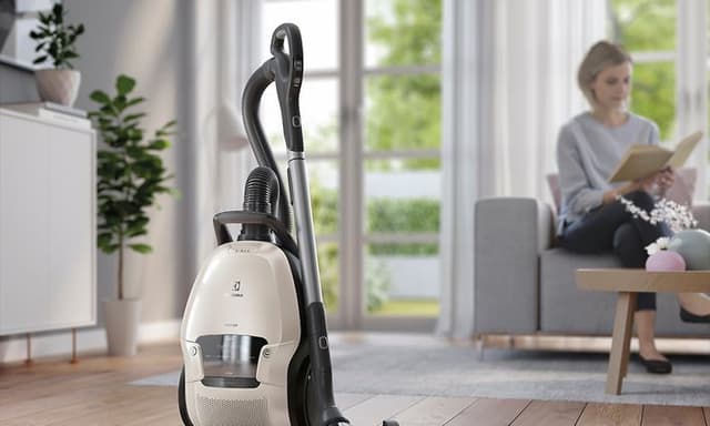 Lys beige Electrolux vanlig støvsuger med skaft som står i en ren stue og en kvinne som leser i bakgrunnen