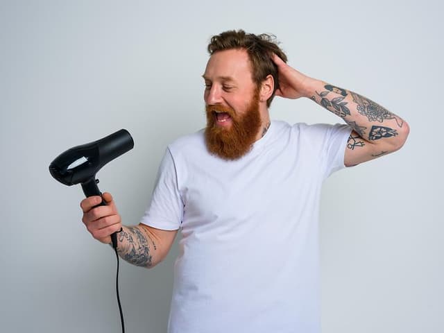 Glad man med vit t-shirt, skägg och tatueringar som sjunger in i en svart hårtork