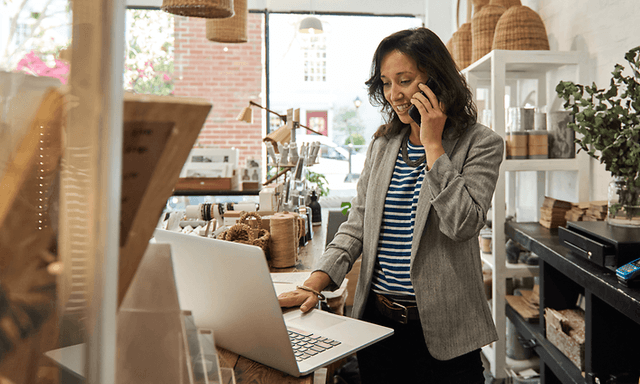 B2B - Elkjøp Control - A woman speaking on her smartphone in front of her laptop in a shop