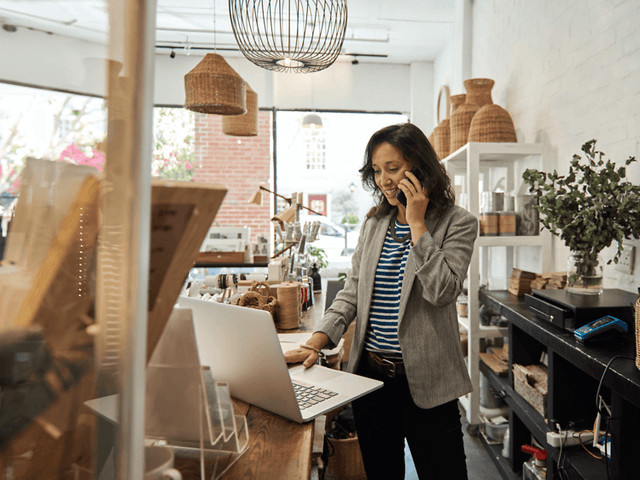 B2B - Elkjøp Control - A woman speaking on her smartphone in front of her laptop in a shop