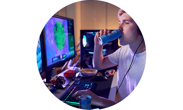 Billede af person der drikker energidrik ved gaming-setup inklusive keyboard, mus og skærm