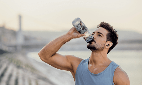 Vältränad man med grått linne och in-ears-hörlurar som står utomhus dricker ur en vattenflaska 