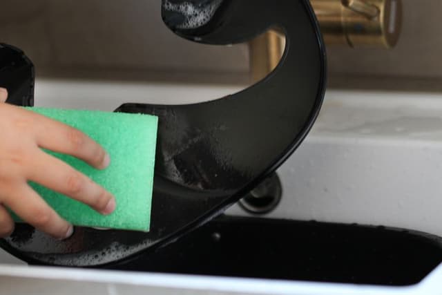 Person som rengör en airfryer med en grön disksvamp i en vit fyrkantig diskho med mässingkran i bakgrunden