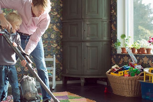 Isä auttaa pientä lastaan imuroimaan hänen huoneensa värikästä mattoa