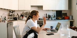 Kvinde der sidder foran en laptop på et spisebord og vinker til webbkameraet