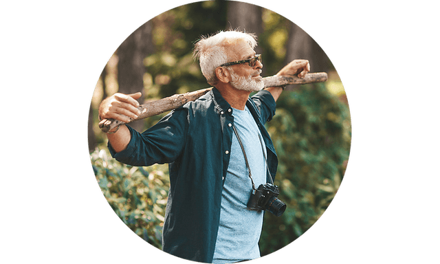 An old man with a camera holds a stick on his shoulders in a forest