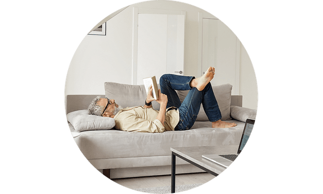 An older man lies on a grey couch reading a book