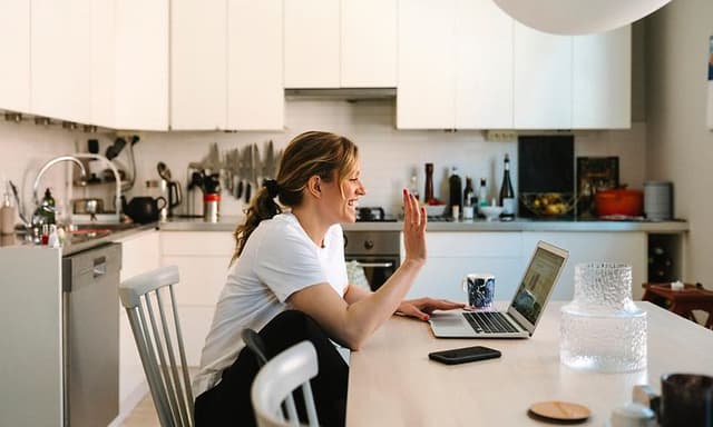 Nainen työskentelee kotona keittiönpöydän ääressä ja vilkuttaa kohti kannettavan näyttöä muille etäkokoukseen osallistujille