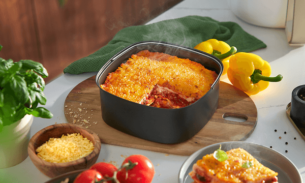 Philips Airfryer's separate baking tray on a table next to a slice of lasagne, fresh vegetables and basil