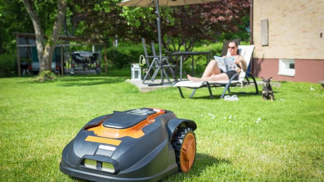 Nainen rentoutuu puutarhassa samalla, kun robottiruohonleikkuri siistii nurmikkoa