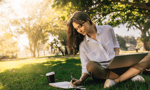 Woman listening music from in-ear-headphones and working on her laptop while sitting at park